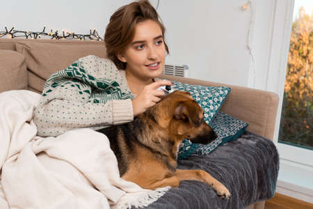 Blond girl with perfect smile, playing video game, enjoying virtual reality, winter, christmas theme, warm tones, cozy vibes, cute small brown mongrel, self isolation time, beige blanketの写真素材