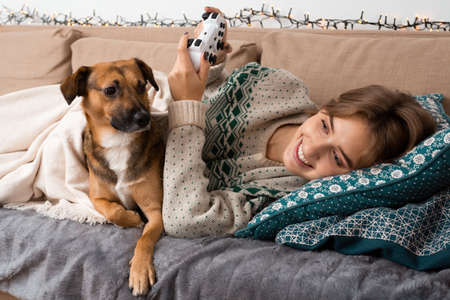 Pretty caucasian blond girl playing video games on a comfy sofa with beige cable knit sweater with her brown cute mongrel. Having fun while self isolation, winter christmas theme, time, cozy vibeの写真素材