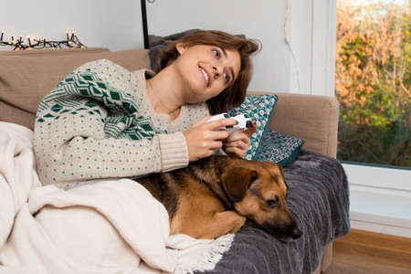 Happy blond woman with smile playing new video game and being immersed fully in amazing big open virtual reality world, warm winter beige cable knit sweater with green pattern, cute brown mongrelの写真素材