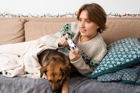 Short haired blond girl playing new video game on her console lying on couch with her dog, being immersed, focused, happy, virtual reality, winter warm sweater, christmas theme, safe home cozy vibe,の写真素材