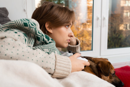 Short haired blond caucasian woman lying on beige sofa with her dog and playing new video games,...