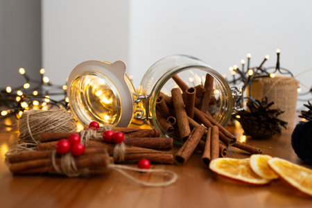 Glass jar with cinnamon sticks falling out on wooden table, with many christmas rustic style, handmade diy ornaments, blurry light in the background, warm tones and winter mood, front viewの写真素材