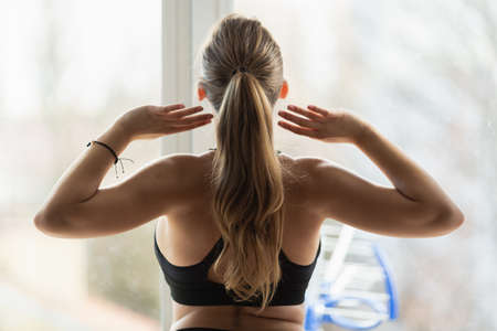 Young blonde caucasian woman stretching in front of the window. Home spring sport exercises. being on diet, Back view. Body positive workout.の写真素材