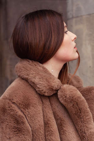 Beautiful brunette pale woman profile wearing brown fur coat with silver earrings. Urban surroundings. Glamor, elegant style. Beige, nude tones. Early spring vibes.の写真素材