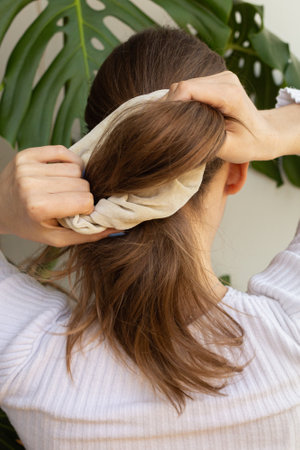 Blonde woman making a ponytail with beige scrunchie, shot from behind, focus on hands, photo in movement. Monstera's leaf in the background.の写真素材