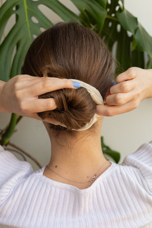 Blonde woman making clean low bun, ballet style with beige scrunchie, focus on slim neck, hands, fingers and nails, view from behindの写真素材