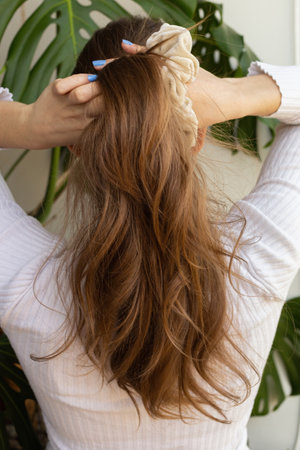 Beautiful long healthy natural blond hair with beige scrunchie view from the back. Caucasian woman styling her hair. Warm positive tones. Monstera plants in the background. Summer vibes.の写真素材