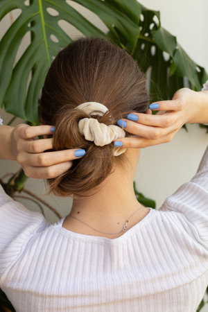 Woman making low messy bun with beige scrunchie against white background wall and green monstera plant with long natural healthy blond hair. Purple nails, silver necklace and white shirt.の写真素材