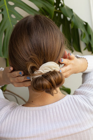 Woman making bun hairstyle with beige scrunchie view from the back against white background and monstera plant. Beautiful blond natural hair.の写真素材