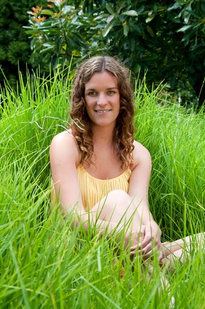 Country girl sitting in long green grass with brown ringlet hair and yellow dress in summerの写真素材