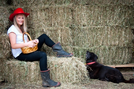 Country girl sitting on hay bale with basket with her faithfull black farm dogの写真素材
