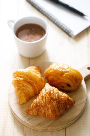A cup of hot chocolate and croissant on wooden background with notebook.  Work from home concept.の写真素材