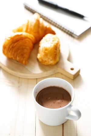 A cup of hot chocolate and croissant on wooden background with notebook.  Work from home concept.の写真素材