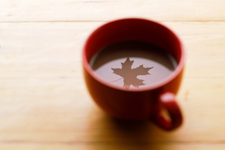 Cup of delicious hot chocolate with red maple leaf reflection on wooden table background. Selective focus.の写真素材