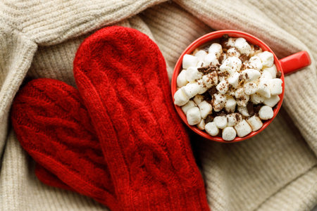 Cup of delicious hot chocolate and marshmallows on warm sweater under warm light with red knitted...
