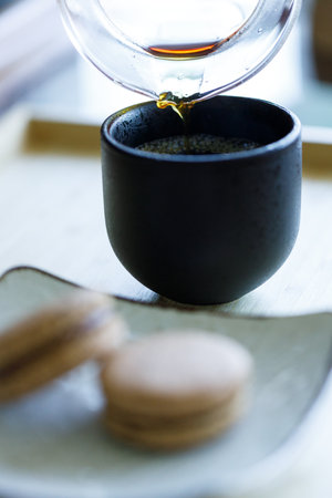 Black coffee pouring into cup and black tea flavor macaron on wooden background. Selective focus.の写真素材
