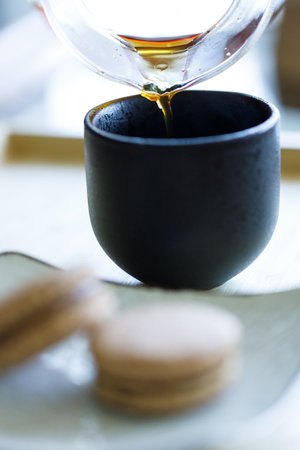 Black coffee pouring into cup and black tea flavor macaron on wooden background. Selective focus.の写真素材