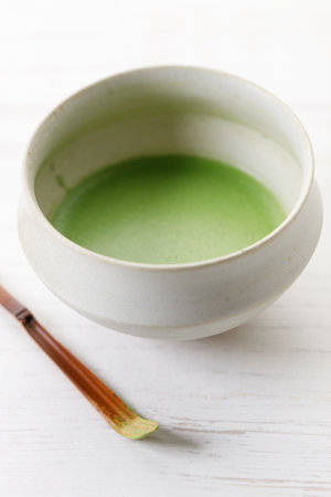 Cup of fresh matcha tea, green tea and Chashaku or tea scoop, equipments for preparing matcha green tea, on white wooden table. Selective focus.の写真素材