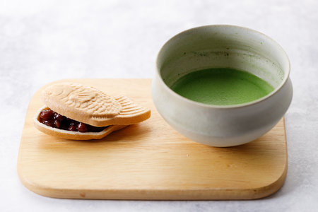 Cup of fresh matcha tea, green tea and Taiyaki, Japanese fish-shaped cake, filled with Anko or red bean on wooden plate on white table. Selective focus.の写真素材