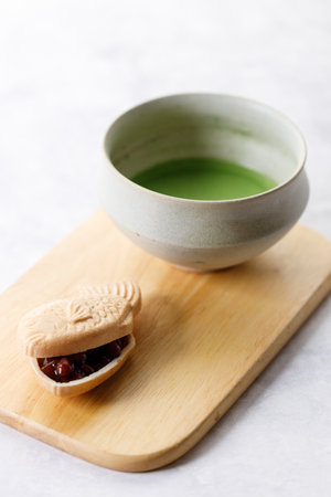 Cup of fresh matcha tea, green tea and Taiyaki, Japanese fish-shaped cake, filled with Anko or red bean on wooden plate on white table. Selective focus.の写真素材