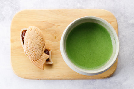 Cup of fresh matcha tea, green tea and Taiyaki, Japanese fish-shaped cake, filled with Anko or red bean on wooden plate on white table. Selective focus.の写真素材