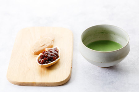 Cup of fresh matcha tea, green tea and Taiyaki, Japanese fish-shaped cake, filled with Anko or red bean on wooden plate on white table. Selective focus.の写真素材