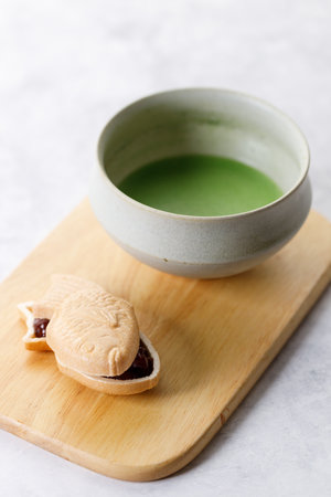 Cup of fresh matcha tea, green tea and Taiyaki, Japanese fish-shaped cake, filled with Anko or red bean on wooden plate on white table. Selective focus.の写真素材