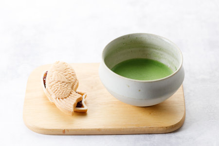 Cup of fresh matcha tea, green tea and Taiyaki, Japanese fish-shaped cake, filled with Anko or red bean on wooden plate on white table. Selective focus.の写真素材