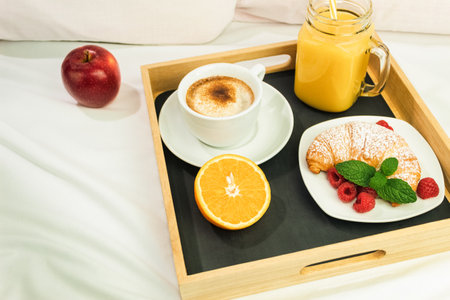 Coffee, orange juice and a croissant on a wooden stalk. Romantic breakfast in bed on white sheets, light from the window. Morning warm light from the window as backgroundの写真素材