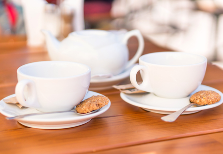 Two white cups of tea and spoons with biscuits, teapot on a wooden tableの写真素材