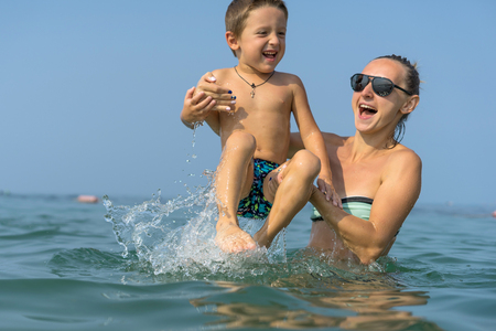 Young mother in black sunglasses and smiling baby boy son in green baseball cap playing in the sea in the day time. Positive human emotions, feelings, joy. Spring and summer holidays.の写真素材