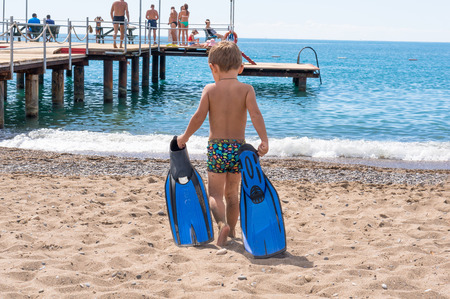 Adorable little blond kid boy having fun on tropical beach. Excited child playing and surfing in sun protected swimsuit in ocean on vacations. White sand, Kid holding flippers for swimmingの写真素材