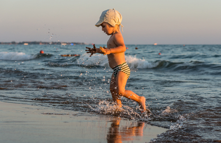 Smiling little baby boy running with splash in the sea on the beach at sunset.の写真素材