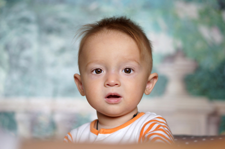 A pretty baby boy in a vest just woken up sitting on a bed with a motherの写真素材