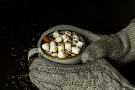 Hands in mittens hold cup of hot cocoa or chocolate with marshmallow on black table from above. Warm...