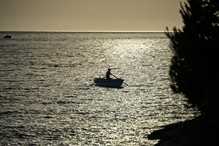 Silhouette of a fisherman in boat on the sea in the evening before sunset with tree silhouette on...