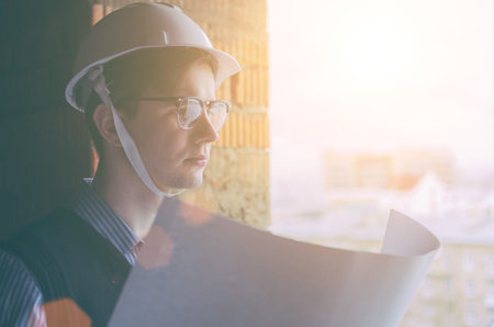 Young man student engineer in glasses and white helmet, with paper in hand in sunset time
