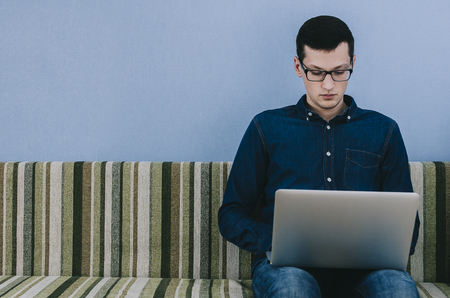 Young man with glasses sitting on sofa with laptopの写真素材
