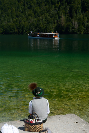 Schonau am Konigssee, Germany - August 30, 2015: Boy wearing Bavaria traditional clothes sitting on lake bank and passer boat. Unidentified people visible.のeditorial素材