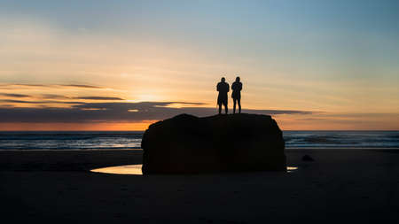 Silhouette image of two people standing on big rock and watching sunset at Muriwai beach, Waitakere, Aucklandの写真素材