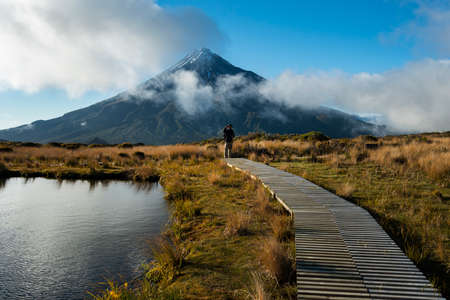 Hiker standing on the wooden boardwalk platform around the Pouakai tarn. Mt Taranaki in the background.の写真素材