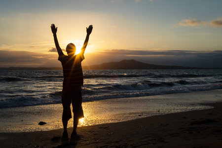 Silhouette of a man with open arms greeting the sun rising over Rangitoto Island, Milford Beach,...