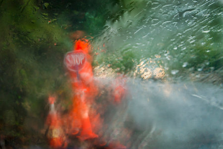 Worker wearing high visibility orange clothing with Stop sign in the heavy rain. Photo taken using intentional camera movement behind rain spattered car window.の写真素材