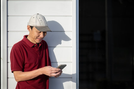 Man smiling while checking his smartphone. Looking at smartphone. Leaning against a shop white wall and glass door.の写真素材