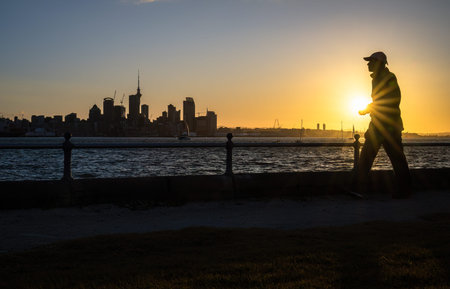 Man walking at Devonport waterfront at sunset. Skytower and Auckland city high-rises in the background.の写真素材