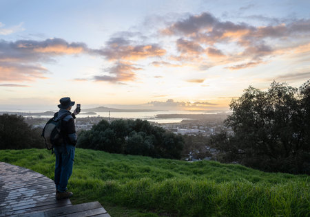 Tourist taking photo using smartphone at Mt Eden summit at sunrise. Rangitoto Island in the distance. Auckland.の写真素材