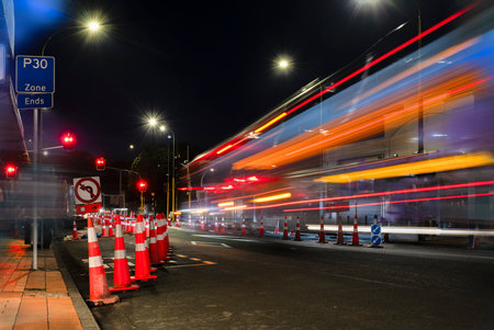 Orange traffic cones lined up on the road. Bus light trails approaching red traffic lights. Auckland.の写真素材