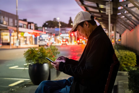 Man waiting for bus while checking his smartphone. Out-of-focus cars in the distance. Auckland.の写真素材