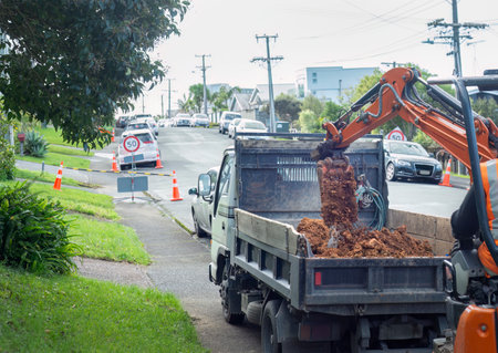 Excavator digging up dirt and moving into truck. Traffic cone and cars along the residential street. Auckland.の写真素材