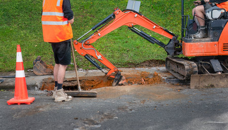 Man driving excavator, digging up dirt between cut concrete pavement. Man with hi-visibility vest holding broom standing by.の写真素材
